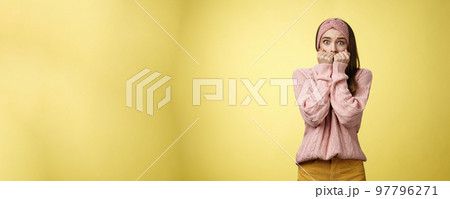 Stunned scared timid insecure cute young girl frightened gasping from fear pressing hands to mouth standing in stupor shocked, horrified stupified against yellow background being victim of abuse 97796271
