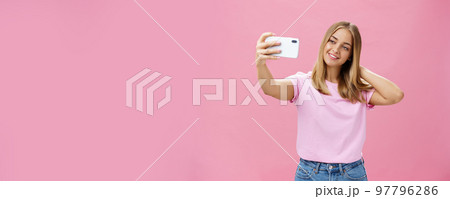 Female beauty blogger taking selfie post new look online. Portrait of charming tanned young woman in t-shirt touching hair gently pulling hand with smartphone near face taking photo over pink wall 97796286