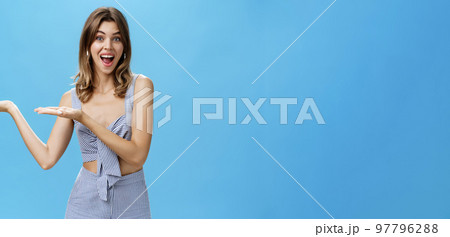 Woman presenting product over copy space. Good-looking charismatic adult tall woman with chestnut hair pointing at left side of wall and smiling broadly as if advertising against blue background 97796288