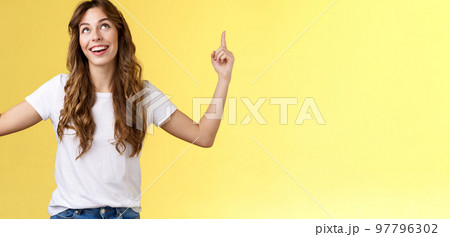Happy admiring cute european curly-haired girl long haircut look pointing up impressed amused smiling broadly satisfactory delighted stand white t-shirt yellow background joyfully react good promo 97796302
