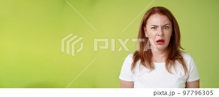 Confused shocked gasping middle-aged redhead woman cringe frustrated puzzled open mouth speechless freak out strange shocking scene stand green background perplexed disappointed 97796305