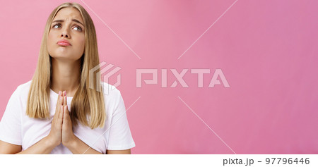 Please make desire become reality. Gloomy upset cute young female student with blond hair and tanned skin without makeup pursing lips sad and lonely holding hands in pray looking up, making wish 97796446