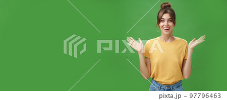Amazed and surprised woman reacting to astonishing gift raising hands near shoulders smiling happily at camera being delighted and pleased with awesome surprise made by friends over green wall 97796463