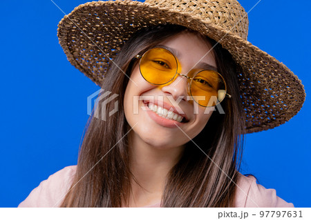 Charming woman in yellow eyewear and straw hat on blue studio backdrop. Smiling positive lady with Charming woman in yellow eyewear and straw hat on blue studio backdrop. Smiling positive lady with 97797631