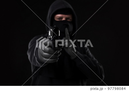Masked robber with gun aiming into the camera against a black background 97798084