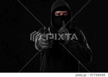 Masked robber with gun aiming into the camera against a black background Masked robber with gun aiming into the camera against a black background 97798086