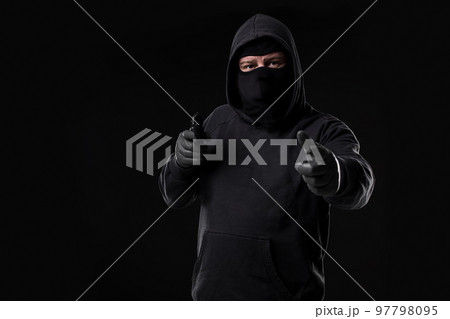 Masked robber with gun aiming into the camera against a black background Masked robber with gun aiming into the camera against a black background 97798095