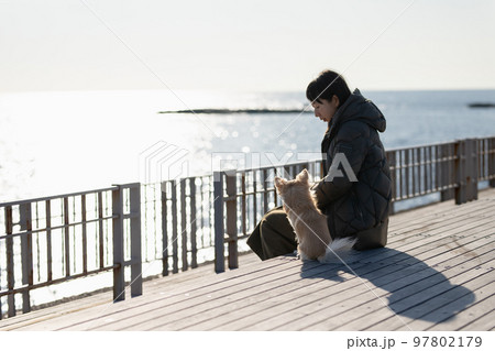 冬の海を背景にしたテラスデッキで愛犬と一緒に寛ぐ飼い主の女性 97802179