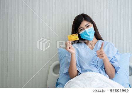 Happy Asian woman wearing a medical mask and holding mock up credit card /insurance card and ok sign in a hospital bed. Insurance policy by bank, payment medical treatment concept 97802960