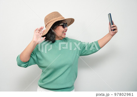 Portrait of happy Asian senior woman wearing sunglasses, a hat, and holding smartphone for a selfie, standing isolated on white background. Concept of tourist enjoy and fun valentine day after retire 97802961