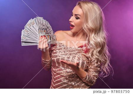 Blonde woman with a perfect hairstyle and bright make-up is posing with fan of hundred dollar bills in her hands. Casino, poker. 97803750