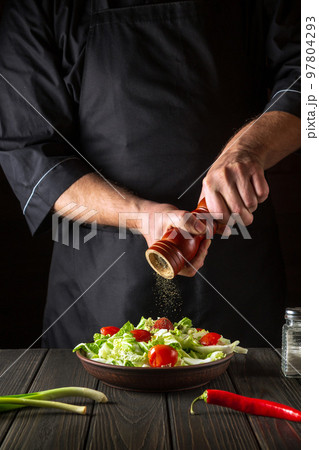 Chef adds peppers to a fresh vegetable salad in a restaurant kitchen. Close-up of cook hands holding a mill. Cooking healthy and tasty food 97804293