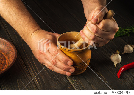 Cook crushing a blending red pepper in a wooden pestle and mortar in a close up view on his hands. Cooking a national dish in the kitchen Cook crushing a blending red pepper in a wooden pestle and mortar in a close up view on his hands. Cooking a national dish in the kitchen 97804298