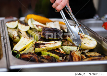 Grilled vegetables on open kitchen food festival event. Street food and outdoor cooking concept. Hand of woman with thongs. Grilled vegetables on open kitchen food festival event. Street food and outdoor cooking concept. Hand of woman with thongs. 97805926
