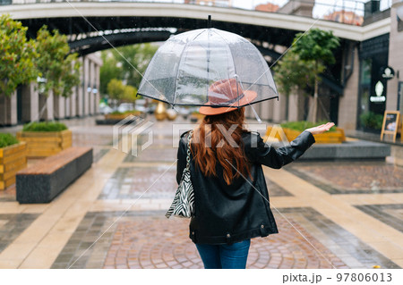 Back view to unrecognizable elegant woman in fashion hat walking on beautiful city street with transparent umbrella, feeling rain with hand, enjoying rainy weather outdoor. Concept of female lifestyle 97806013