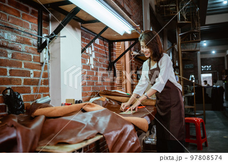 Tailor in apron working happily with leather in leather workshop Tailor in apron working happily with leather in leather workshop 97808574