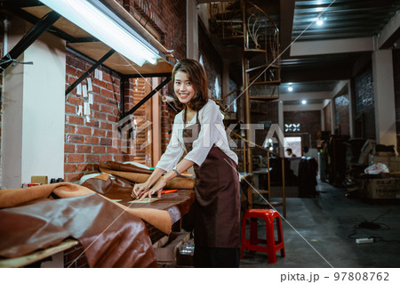 Tailor in apron smiling at camera while measuring leather material 97808762