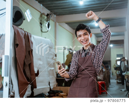 excited asian craftsman smiling using smartphone at leather craft workshop excited asian craftsman smiling using smartphone at leather craft workshop 97808775