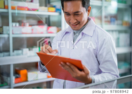 Asian male pharmacist in uniform working with clipboard while registering Asian male pharmacist in uniform working with clipboard while registering 97808942