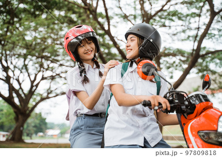 Two student girls wearing helmets chatting while riding a motorcycle Two student girls wearing helmets chatting while riding a motorcycle 97809818