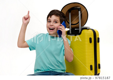 Teenage traveler boy in blue turquoise t-shirt, with yellow suitcase, talking on mobile phone, pointing forefinger at copy space on white background, looking at camera, expressing positive emotions 97815187