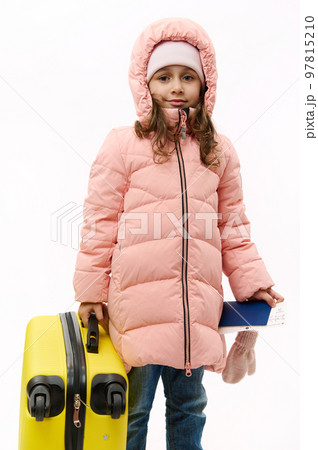 Adorable Caucasian baby girl wearing pink down coat, going for winter holidays, travelling abroad, holding boarding pass and yellow suitcase, smiling looking at camera over white backdrop. Copy space 97815210