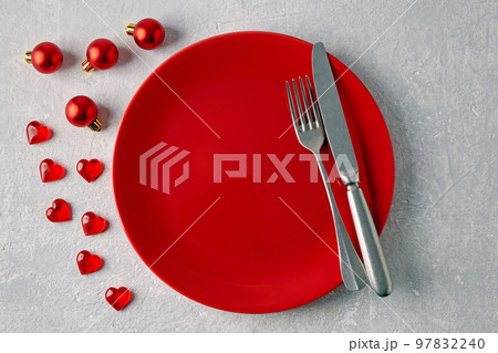 An empty red ceramic plate with cutlery surrounded by red christmas balls and hearts on a light concrete table 97832240