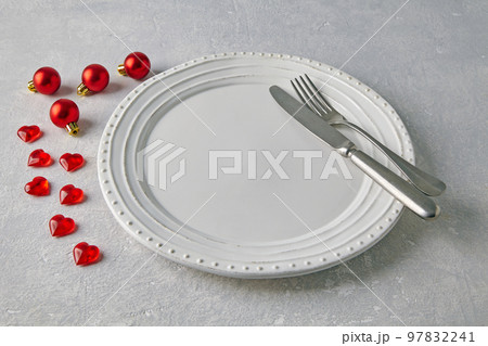 An empty white ceramic plate with cutlery surrounded by red christmas balls and hearts on a light concrete table 97832241