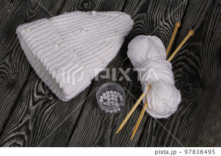 Knitted scarf in white. Women's handmade knitted clothes. Nearby is a skein of white thread for knitting and knitting needles. On black boards. Knitted scarf in white. Women's handmade knitted clothes. Nearby is a skein of white thread for knitting and knitting needles. On black boards. 97836495