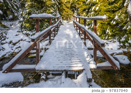 Small wooden footbridge covered by snow over icy mountain creek in wintertime. 97837040