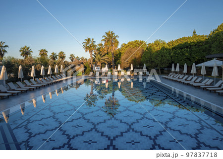 Empty loungers and folded parasols surrounded with trees arranged at poolside with clear blue sky in the background at tourist resort Empty loungers and folded parasols surrounded with trees arranged at poolside with clear blue sky in the background at tourist resort 97837718
