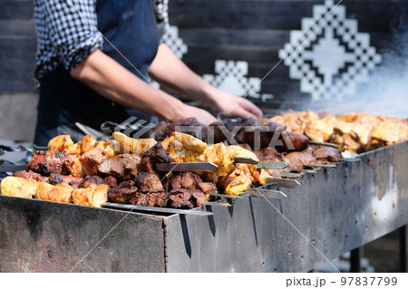 Tasty fried pieces of meat on skewers are roasted on grill at festival outdoor. Chef in black apron prepares barbecue. Shish kebab national cuisine of Central Asia. Tasty fried pieces of meat on skewers are roasted on grill at festival outdoor. Chef in black apron prepares barbecue. Shish kebab national cuisine of Central Asia. 97837799