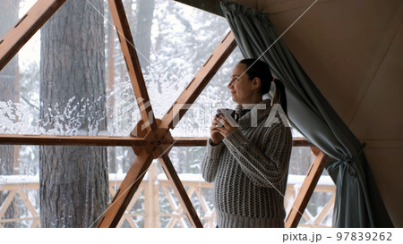 Young caucasian woman drinking hot chocolate or tea over panoramic window of dome camping, enjoying amazing view of snow-covered nature Young caucasian woman drinking hot chocolate or tea over panoramic window of dome camping, enjoying amazing view of snow-covered nature 97839262