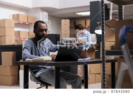 African american employee sitting at desk in warehouse delivery department looking at cargo stock on laptop before start preparing helmet order. Storehouse worker using carton box and bubble wrap African american employee sitting at desk in warehouse delivery department looking at cargo stock on laptop before start preparing helmet order. Storehouse worker using carton box and bubble wrap 97843080