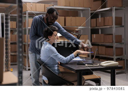 Warehouse employees analyzing delivery checklist on laptop computer searching for metallic box order before shipping package. Multi ethnic team analyzing at transportation logistics while working in 97843081