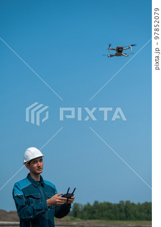 A man in a helmet and overalls controls a drone at a construction site. The builder carries out technical oversight. 97852079