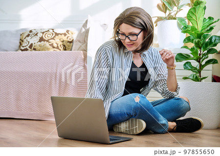 Mature woman sitting on the floor at home looking at laptop Mature woman sitting on the floor at home looking at laptop 97855650