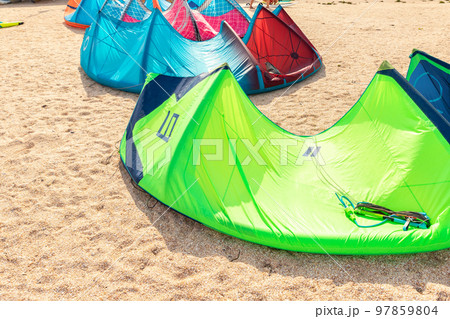 Close-up many bright kitesurf wings and kite equipment sand beach shore watersport spot on bright sunny day against sea coast blue sky and kiter riding surf school camp. Active travel sport concept 97859804