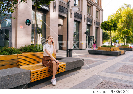 Wide shot of elegant young woman with blond hair having mobile phone conversation sitting on bench on city street in summer day. Focused lady making call on mobile phone, speaking about work issues. Wide shot of elegant young woman with blond hair having mobile phone conversation sitting on bench on city street in summer day. Focused lady making call on mobile phone, speaking about work issues. 97860070