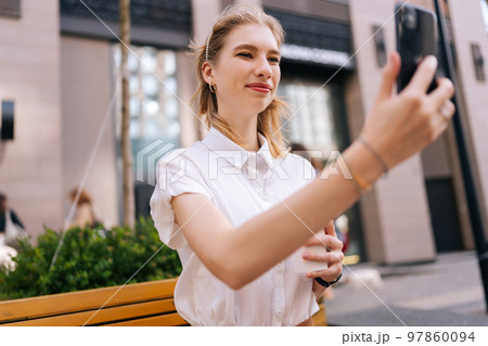 Low-angle view of pretty young woman taking selfie using smartphone and holding cup with takeaway coffee in summer day sitting on bench on city street. Cute lady having video call via mobile phone. 97860094