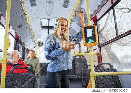 Portrait of fair long haired female wearing blue sweater and black trousers with earmuffs standing near bus payment terminal and performing contactless operation passengers watching city from window. Portrait of fair long haired female wearing blue sweater and black trousers with earmuffs standing near bus payment terminal and performing contactless operation passengers watching city from window. 97860145
