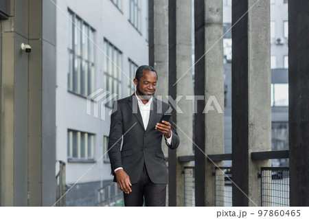 Happy successful African American businessman boss walking outside office building, manager holding smartphone reading message and smiling, using communication app. Happy successful African American businessman boss walking outside office building, manager holding smartphone reading message and smiling, using communication app. 97860465