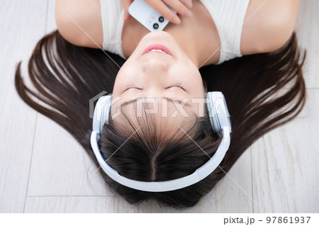 フローリングに寝転んでスマホ音楽に浸る女性 97861937