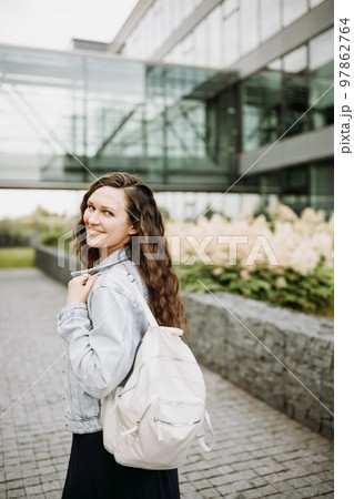 Happy pretty young brunette woman with white nylon backpack in blue denim jacket outdoor. Close up, urban street portrait Happy pretty young brunette woman with white nylon backpack in blue denim jacket outdoor. Close up, urban street portrait 97862764