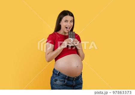 Portrait Of Cheerful Young Pregnant Lady Biting Chocolate Bar Over Yellow Background Portrait Of Cheerful Young Pregnant Lady Biting Chocolate Bar Over Yellow Background 97863373
