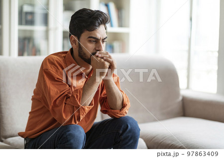 Pensive young middle eastern man sitting on sofa at home Pensive young middle eastern man sitting on sofa at home 97863409
