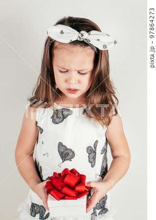 Upset little girl hold beautiful present box with bow looking down. Portrait of child in festive dress on white. 97864273