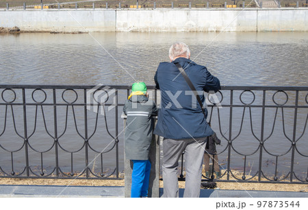A gray-haired grandfather and his grandson are standing on the river embankment and leaning on metal handrails, looking at a fisherman on a sunny spring day 97873544