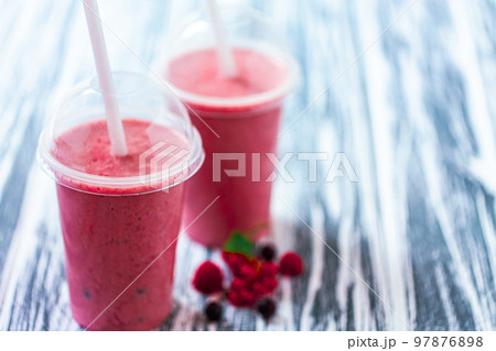 Front view of two berries milkshakes in plastic cups with ingredients on wood table. Space for text Front view of two berries milkshakes in plastic cups with ingredients on wood table. Space for text 97876898