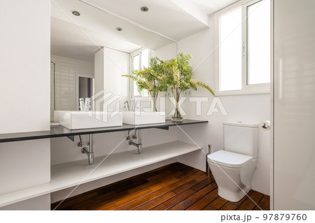 Two square sinks on black marble countertop in spacious bathroom with window to street. Large mirror reflects back of room with white ceramic tiles on wall. Floor is covered with parquet board. 97879760